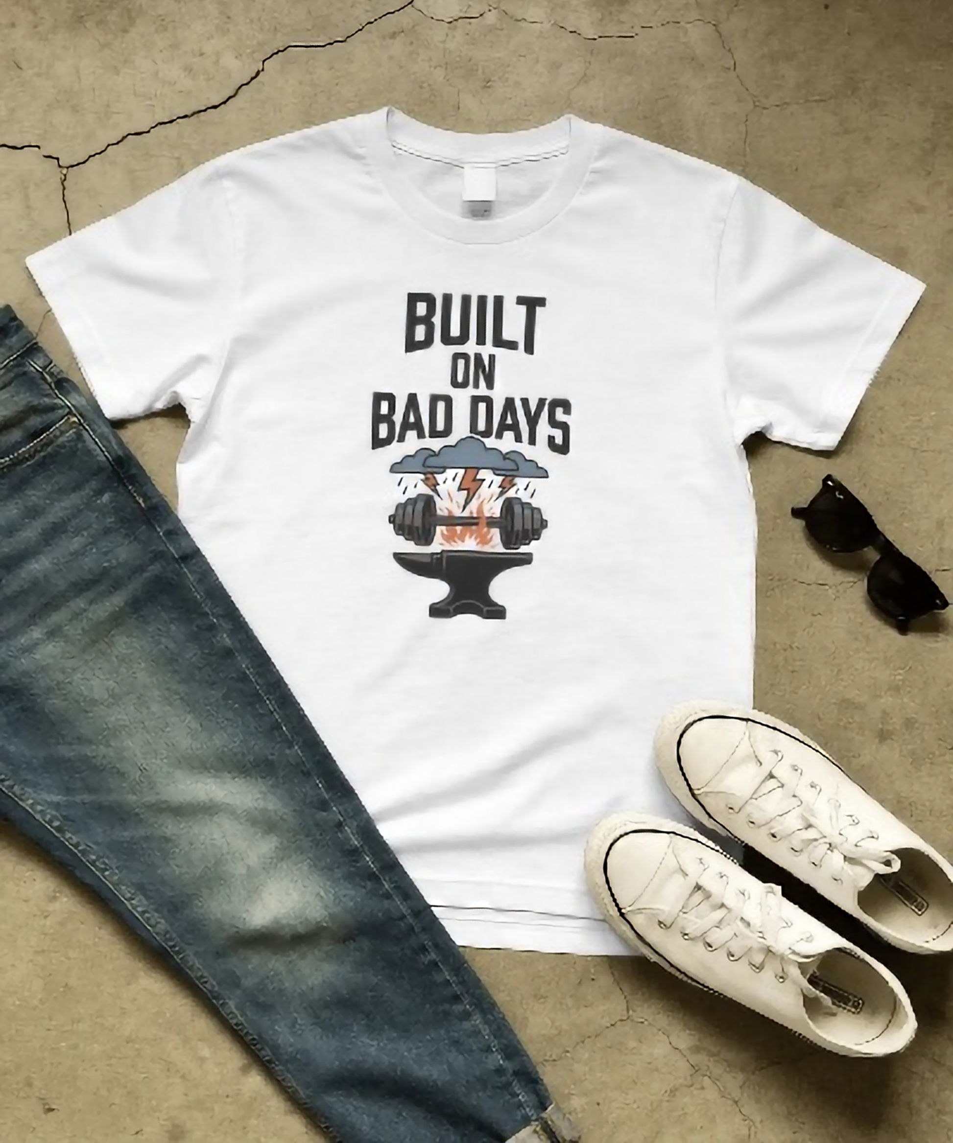 White premium oversized unisex t-shirt by Nite Stream featuring a gritty motivational graphic of a dumbbell on an anvil with lightning and storm clouds, and bold typography reading 'BUILT ON BAD DAYS'.