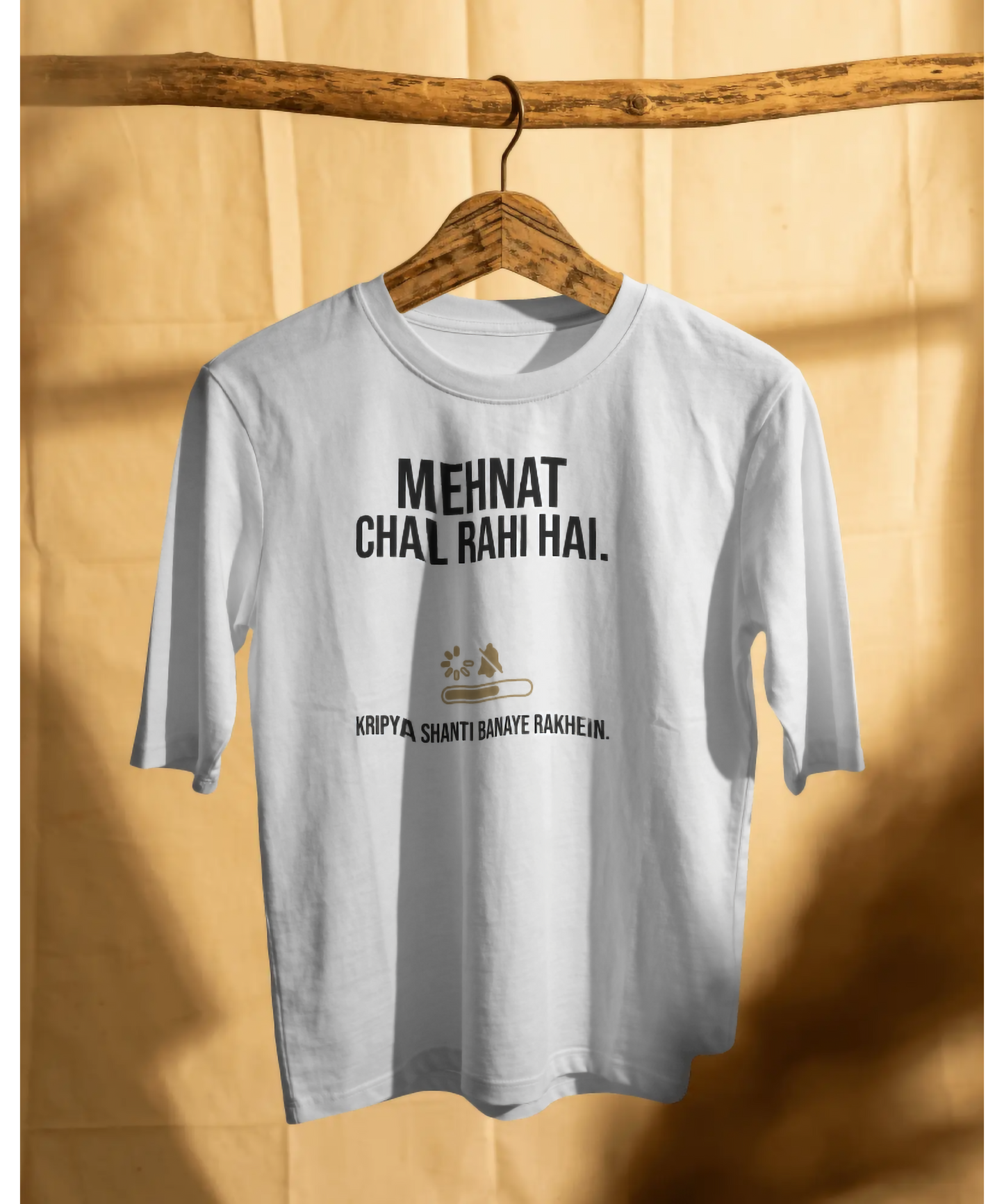 White unisex graphic t-shirt hanging on a wooden branch with warm sunlight and shadows. The t-shirt features a bold typography design saying "Mehnat Chal Rahi Hai" and "Kripya Shanti Banaye Rakhein" with a loading bar and mute icon. Premium hustle culture streetwear by The Nite Stream.
