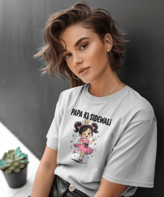 A smiling woman modeling a white graphic t-shirt. The t-shirt features the quirky Hindi text "PAPA KI SIDEWALI" above a cute, vibrant illustration of a happy little girl in a pink dress, winking and pointing. Premium funny desi streetwear by Nite Stream.